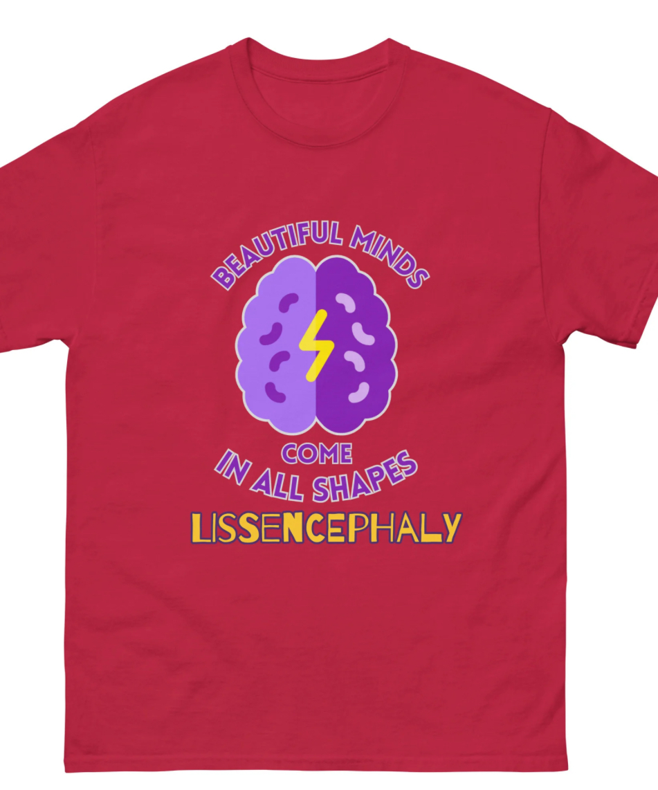 Red tshirt that has a purple brain shaped icon center chest with a diagonal z that looks like a lightning bolt and has arched writing above saying Beautiful Minds and horizontal writing under that says Come with arched writing under sayin In All Shapes with horizontal writing under saying Lissencephaly